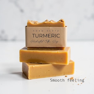 Op de afbeelding: Drie stukken kurkuma zeep op elkaar gestapeld. De bovenste zeep heeft een bruin papieren etiket met de tekst "soap story TURMERIC Handcrafted Tallow Soap". De onderkant van de afbeelding luidt "Smooth feeling".