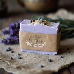 Op de afbeelding: Een rechthoekige zeep in lavendelkleur met een bruine papieren wikkel met de tekst "Imperial Garden". De zeep is bedekt met kleine, donkerblauwe bessen en bruine korrels. Een takje lavendel is zichtbaar op de achtergrond.