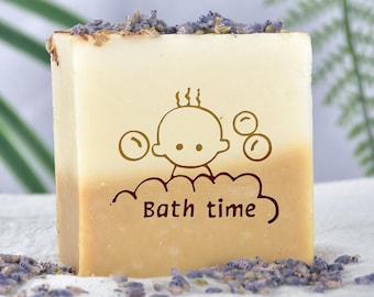 May include: A bar of soap with a light brown and white color scheme. The soap has a design of a baby in a bubble bath with the text "Bath time" printed on it. The soap is surrounded by dried lavender flowers.
