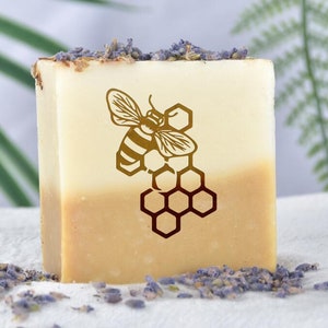 May include: A bar of soap with a honeybee and honeycomb design. The soap is a light yellow color and has a white layer on top. The soap is surrounded by dried lavender flowers.