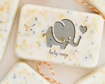 May include: Five bars of handmade soap with a light yellow and white color. Each bar has a small elephant design with the words "baby soap" printed on it. The bars are tied with orange and white twine.