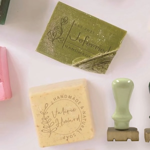 May include: Overhead shot of handmade soap bars in various colors, including green, pink, and beige. The bars are stamped with botanical designs and text, such as "Jeferried Handmade" and "Valerie Howard Soap." Two green soap stamps are also visible.