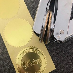 以下が含まれることがあります： 丸いエンボス加工が施された金色の金属製スタンプ。スタンプは、紙やその他の素材に印鑑を押すために使用されます。スタンプは、2つの金色のステッカーが貼られた紙の上に置かれています。ステッカーはギザギザの縁と空白の中央を持っています。1つのステッカーには、「GENUINE ART」と「Osborne」の文字が印刷されています。