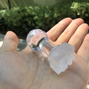 May include: A clear glass stamp with a faceted top and a square base. The base has a raised design.