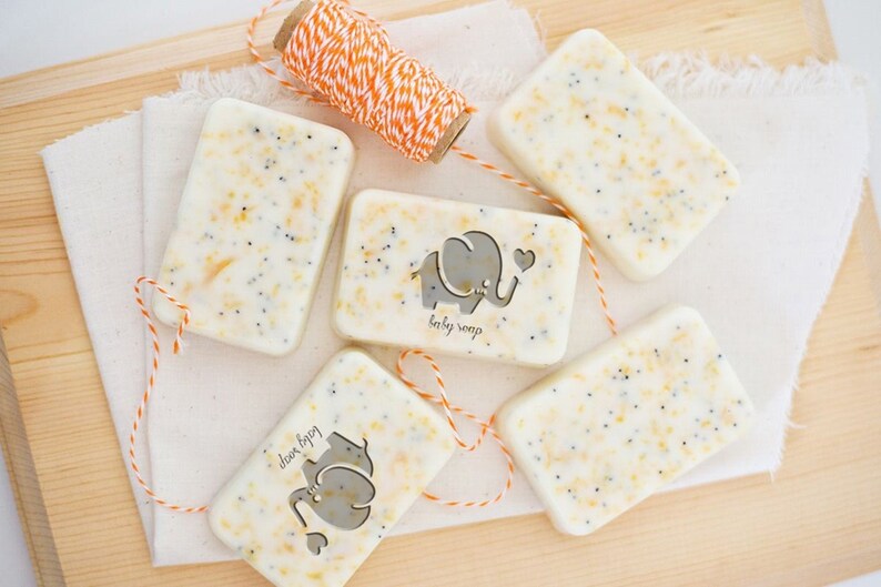 May include: Five bars of handmade soap with a light yellow and white color. Each bar has a small elephant design with the words "baby soap" printed on it. The bars are tied with orange and white twine.