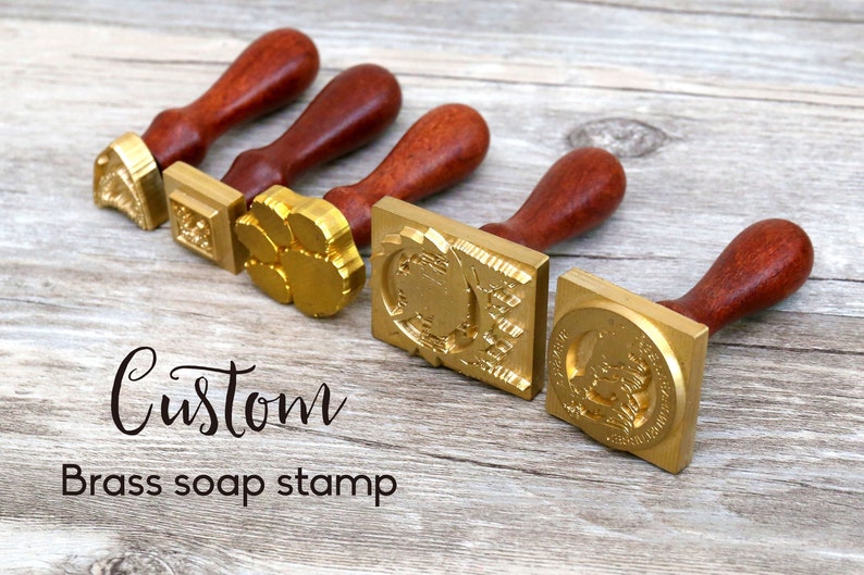 May include: Five brass soap stamps with wooden handles. The stamps feature different designs, including a paw print, a geometric pattern, and a floral design. The text "Custom Brass soap stamp" is visible below the stamps.