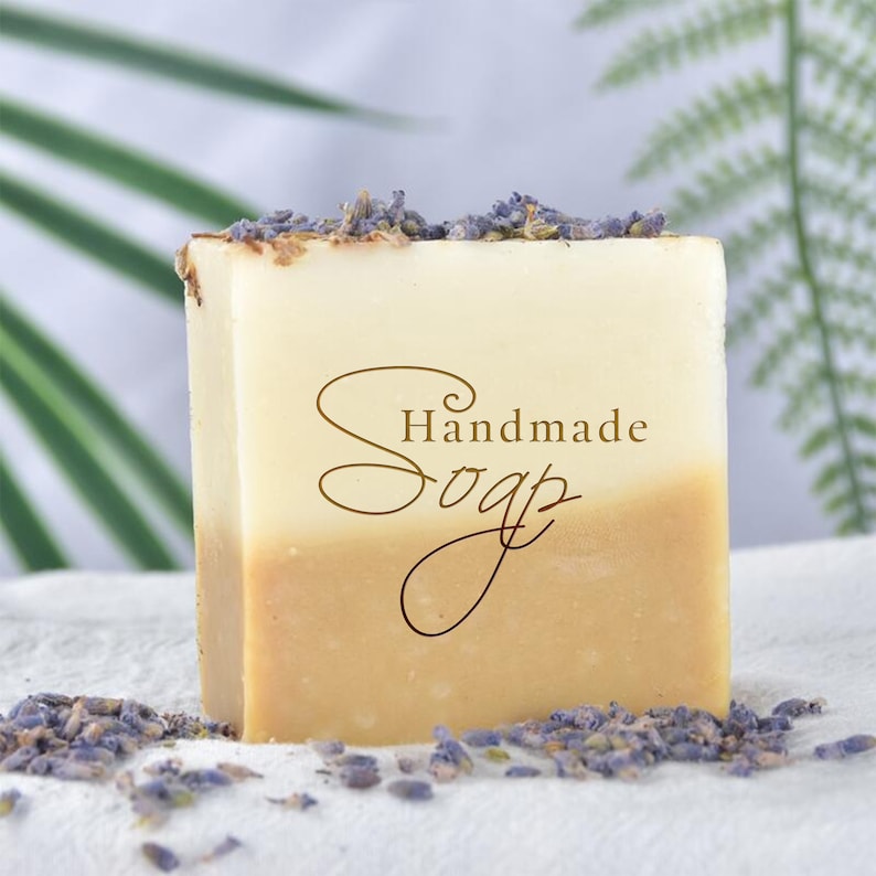 May include: A bar of handmade soap with a light brown and white color. The soap is topped with dried lavender flowers and has the words "Handmade Soap" written on it.