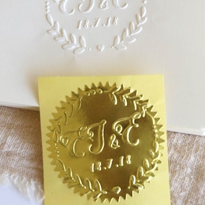 以下が含まれることがあります： 「J&C」のイニシャルと「18.7.18」の日付が書かれた、花輪のデザインの金箔ステッカーです。