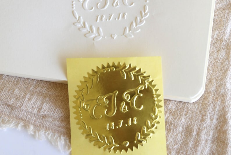 以下が含まれることがあります： 「J&C」のイニシャルと「18.7.18」の日付が書かれた、花輪のデザインの金箔ステッカーです。