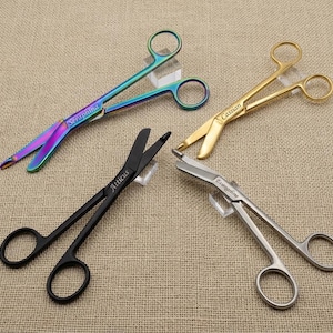 May include: Four pairs of medical trauma shears with finger loops, each featuring a unique finish and engraved name. The colors include iridescent rainbow, gold, matte black, and silver. The names Seraphina, Cassian, Atticus, and Evangeline are engraved.
