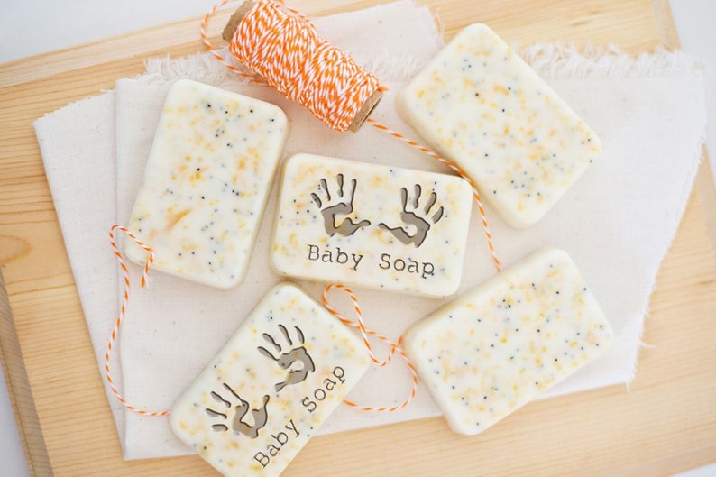 Puede incluir: Cuatro barras de jab&oacute;n blanco con un aspecto moteado de color amarillo claro y gris. Cada barra tiene un dise&ntilde;o de huella de mano y el texto "Baby Soap" impreso en ella. Las barras est&aacute;n atadas con un cordel naranja y blanco.