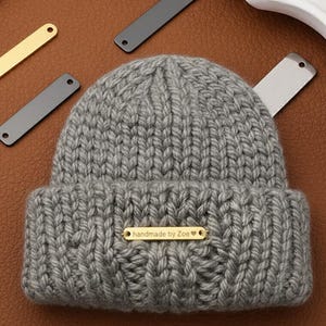 Puede incluir: Un gorro de punto gris con una etiqueta dorada que dice "handmade by Zoe". El gorro está hecho de hilo grueso y tiene un borde doblado. Varias etiquetas metálicas doradas y grises están esparcidas alrededor del gorro.