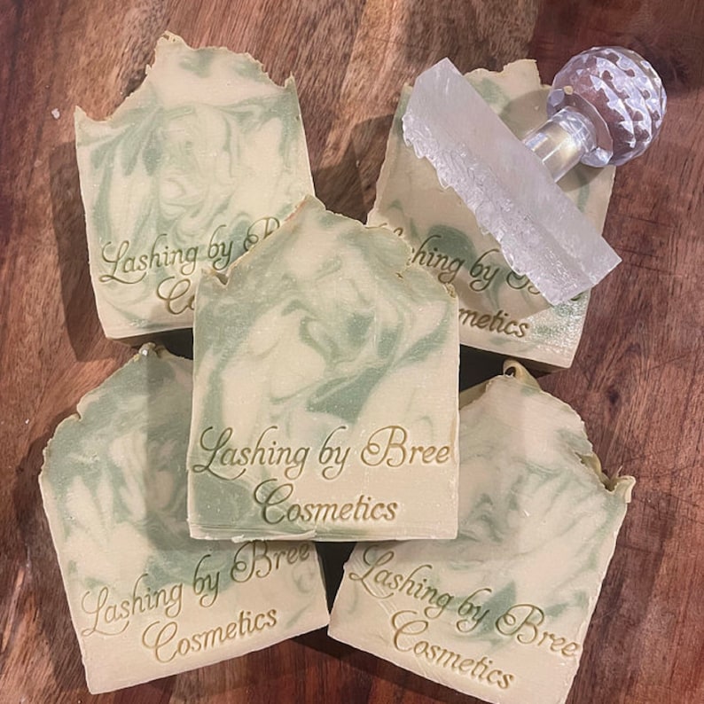 May include: Five handmade soaps with a green and white swirl design, stamped with "Lashing by Bree Cosmetics." A clear, textured soap cutter with a decorative handle rests on top. The soaps are arranged on a wooden surface.