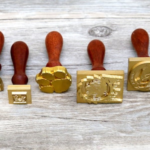 May include: Five wooden-handled wax seal stamps with different designs. The stamps are made of brass and have a gold finish. The designs include a paw print, a geometric pattern, a building, and a crest.