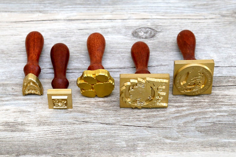 May include: Five wooden-handled wax seal stamps with different designs. The stamps are made of brass and have a gold finish. The designs include a paw print, a geometric pattern, a building, and a crest.