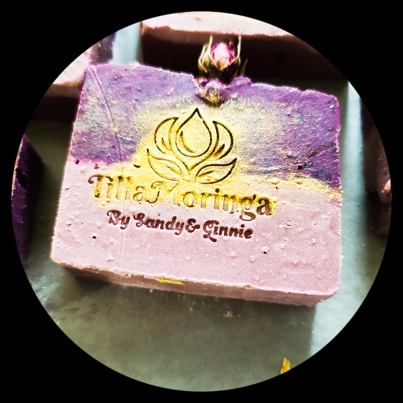 May include: A square bar of handmade soap with a two-tone color scheme of pink and purple, accented with gold flecks. The soap is embossed with a floral design and the text "TillaMoringa By Sandy & Ginnie". A small dried flower bud is attached.