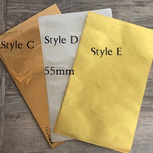 以下が含まれることがあります： 金、白、黄色の異なるデザインのステッカー用紙3枚。シートには「Style C」、「Style D」、「Style E」とラベル付けされています。白いシートには「55mm」の文字が印刷されています。