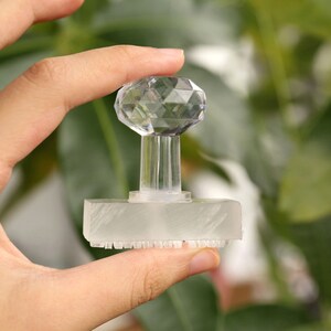 May include: A clear, faceted crystal stamp with a rectangular base and a decorative, geometric top. The stamp is held in a hand against a blurred green background.