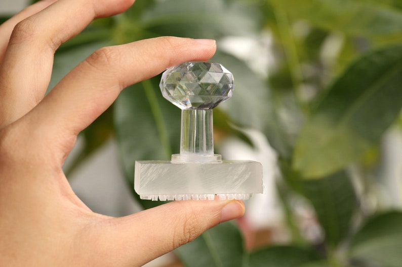 May include: A clear, faceted crystal stamp with a rectangular base and a decorative, geometric top. The stamp is held in a hand against a blurred green background.