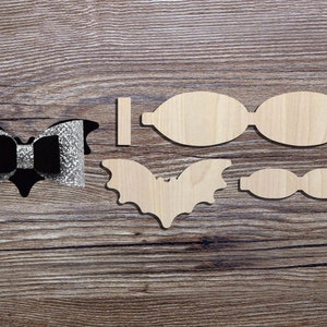 Puede incluir: Un conjunto de suministros de artesanía de madera para hacer un lazo para el pelo con forma de murciélago. El conjunto incluye dos formas de alas de murciélago, dos formas de lazo y una pieza central. El lazo está decorado con purpurina negra y plateada.