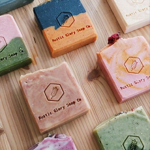 May include: An overhead shot of several handmade soaps in various colors, including pink, green, and orange. Each soap bar has a unique design and is stamped with the text "Rustic Glory Soap Co." on a wooden surface.
