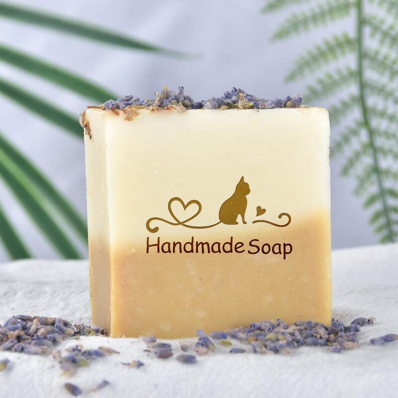 May include: A bar of handmade soap with a light brown base and a white top. The soap has a cat silhouette and a heart design with the text "Handmade Soap" printed on it. The soap is surrounded by dried lavender flowers.