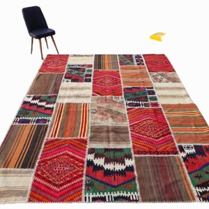 May include: A large, rectangular patchwork rug with a variety of colors and patterns. The rug features geometric designs, stripes, and floral motifs in shades of red, orange, brown, green, and cream. A dark blue chair with wooden legs is in the background.