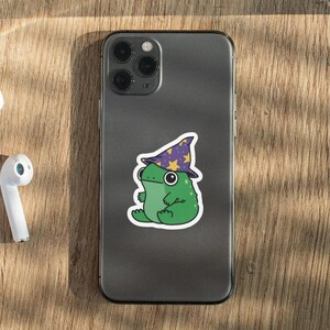 May include: A gray iPhone with a green frog wearing a purple and yellow wizard hat sticker on the back.