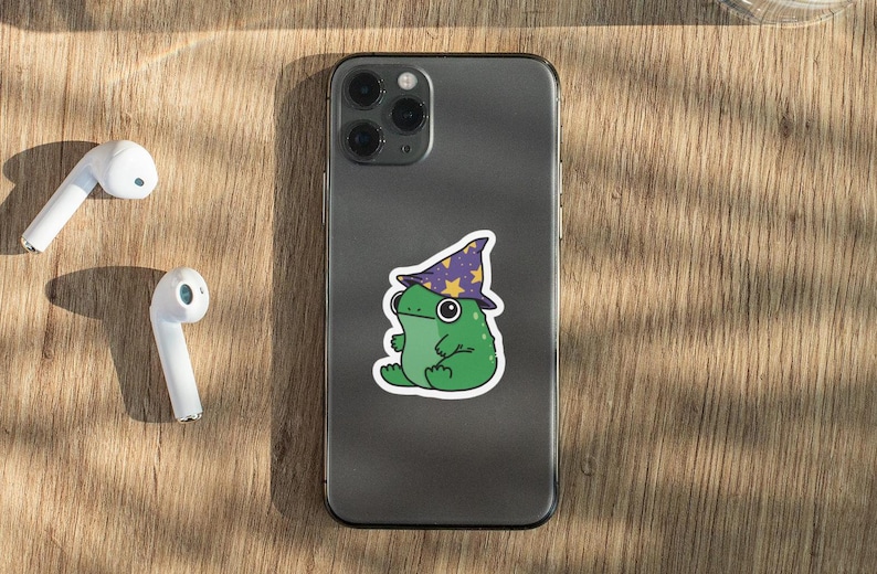May include: A gray iPhone with a green frog wearing a purple and yellow wizard hat sticker on the back.