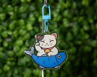 May include: A clear acrylic keychain featuring a cartoon Maneki-neko cat with a raised paw, sitting on a blue fish. The cat has a white body, gray spot, and a red collar with a gold bell. The keychain has a light blue clasp.