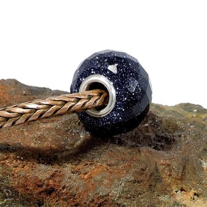 Può includere: Una perla sfaccettata, blu scuro con dettagli color argento, cosparsa di minuscole macchie bianche, è infilata su una catena intrecciata color rame. La perla poggia su una pietra ruvida, strutturata, marrone e verde.