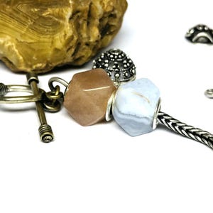 Könnte beinhalten: Nahaufnahme von Schmuckkomponenten, darunter eine facettierte, pfirsichfarbene Edelsteinperle und eine hellblaue Edelsteinperle mit silbernen Akzenten. Eine Silberkette und dekorative Metallperlen sind ebenfalls sichtbar. Der Hintergrund ist weiß.