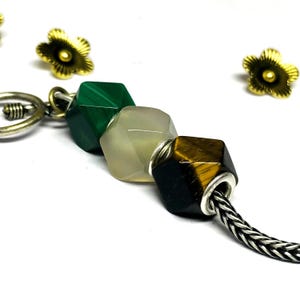 Könnte beinhalten: Nahaufnahme eines Armbands mit drei facettierten Edelsteinperlen in Grün, Creme und Braun. Die Perlen sind an einer silbernen Kette aufgereiht. Zwei goldfarbene, blütenförmige Anhänger befinden sich im Hintergrund.