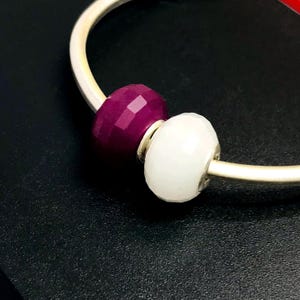 Puede incluir: Una pulsera de plata con dos cuentas facetadas. Una cuenta es de color magenta intenso y la otra blanca. Las cuentas tienen un diseño geométrico multifacético. La pulsera está sobre una superficie negra.
