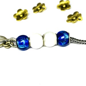 Könnte beinhalten: Ein silberner Schlüsselanhänger mit einer Kette und einer Reihe von Perlen. Die Perlen umfassen zwei weiße Perlen und zwei blaue Glasperlen. Mehrere goldfarbene, blütenförmige Charms sind um die Perlen herum verstreut.