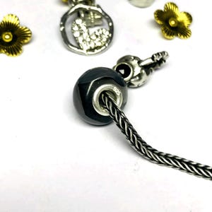 May include: A faceted, dark gray bead with a silver-tone core is strung on a braided silver-tone chain. Other charms include a silver-tone heart pendant, a guitar charm, and gold-tone flower charms. The bead is marked with "925".