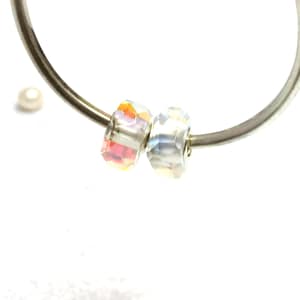 May include: Two faceted crystal beads with iridescent color, one red and orange, the other clear, strung on a silver metal hoop. A small, white pearl is visible in the background. Jewelry.