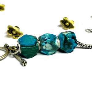Könnte beinhalten: Nahaufnahme einer handgefertigten Halskette mit drei facettierten, türkisfarbenen und grünen Edelsteinperlen. Die Perlen sind an einer Silberkette aufgereiht und mit silbernen Metallringen akzentuiert. Goldfarbene, blütenförmige Perlen sind um die Halskette verstreut.