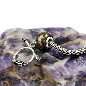 May include: A silver charm bracelet with a brown and white bead and a silver toggle clasp. The bracelet is on a purple and white rock.