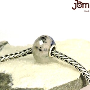 K&ouml;nnte beinhalten: Eine facettierte, ovale Perle mit hellgrauer Basis und dunkelblauen und schwarzen Einschl&uuml;ssen. Die Perle ist an einer silbernen Kette aufgereiht. Das Bild enth&auml;lt den Text "JBM Beads".