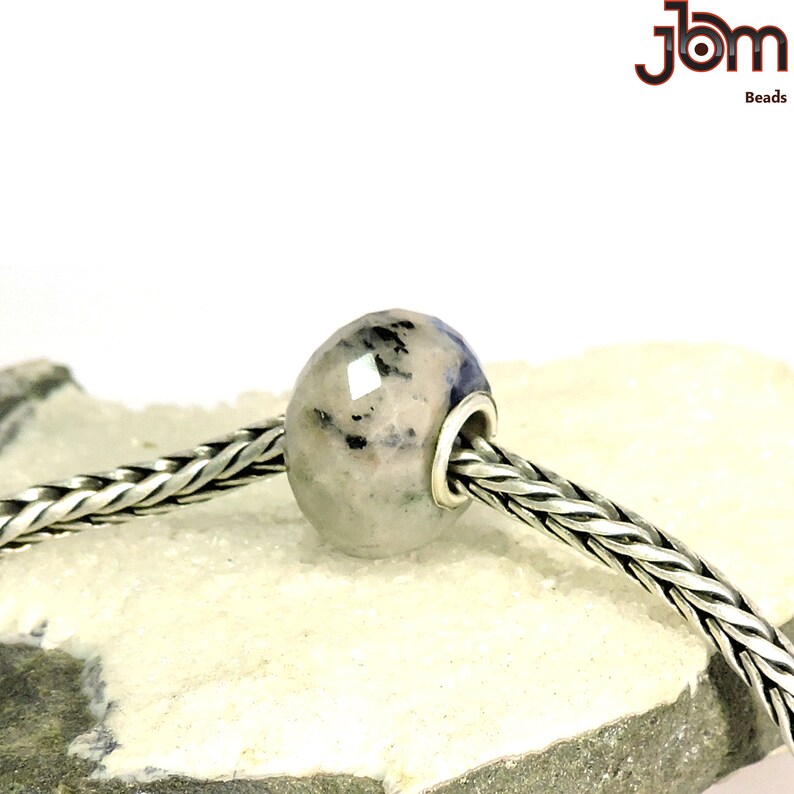 K&ouml;nnte beinhalten: Eine facettierte, ovale Perle mit hellgrauer Basis und dunkelblauen und schwarzen Einschl&uuml;ssen. Die Perle ist an einer silbernen Kette aufgereiht. Das Bild enth&auml;lt den Text "JBM Beads".