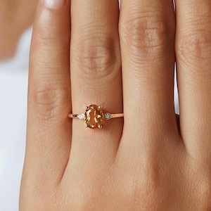 Könnte beinhalten: Ein Goldring mit einem zentralen, oval geschliffenen orangefarbenen Edelstein, der von zwei kleinen, klaren, marquiseförmigen Steinen auf einer dünnen, polierten Goldschiene flankiert wird. Das Design betont die warme Farbe des Steins.