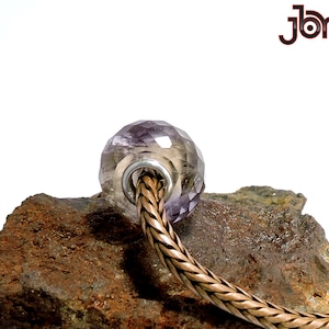 Könnte beinhalten: Eine facettierte Amethyst-Glasperle mit einem silbernen Kern, die auf einem braunen Stein liegt. Die Perle ist Teil eines geflochtenen Kupferarmbands.