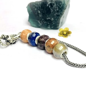 Könnte beinhalten: Ein Armband mit einer Auswahl an farbigen Edelsteinperlen, darunter orange, blau und braun, auf einer Silberkette aufgereiht. Ein Silberverschluss und eine goldene Blume akzentuieren das Design. Ein großer, grüner Stein befindet sich im Hintergrund.