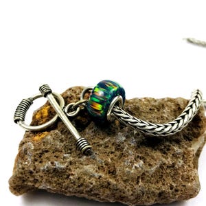 May include: A silver toggle clasp and a bead with a dark blue base and iridescent stripes of green, yellow, and orange. The bead is strung on a silver chain. The clasp and bead are resting on a rough, brown stone.