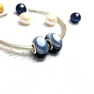 Puede incluir: Una pulsera de plata con dos cuentas de vidrio azul y blanco con un diseño a rayas. Varias perlas en tonos blanco, dorado y azul marino están dispersas alrededor de la pulsera.