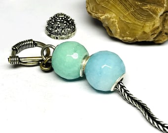 Puede incluir: Primer plano de una pieza de joyería con dos cuentas redondas facetadas, azul claro y verde agua. Las cuentas están conectadas a una cadena y un cierre de plata. Elementos decorativos plateados adicionales están dispersos.