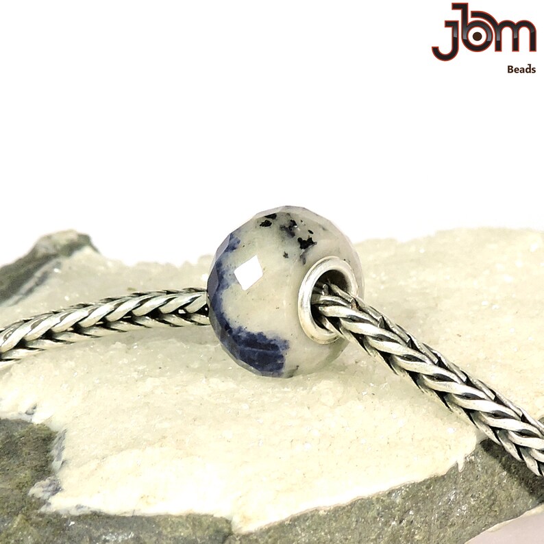 K&ouml;nnte beinhalten: Eine facettierte Perle mit einem hellgrauen und blauen Muster, auf einer silbernen Kette aufgef&auml;delt. Die Perle hat einen silberfarbenen Metallkern. Das Bild enth&auml;lt den Text "JBM Beads".