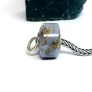 Könnte beinhalten: Eine zylindrische Perle mit glatter, polierter Oberfläche in einem sanften Kornblumenblau, die mit verstreuten goldbraunen Einschlüssen versehen ist. Die Perle ist auf eine Silberkette gefädelt und hat einen kleinen, runden Silberring an der Seite.