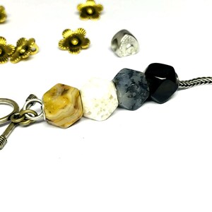 Könnte beinhalten: Nahaufnahme eines Strangs aus Edelsteinperlen in Gelb-, Weiß-, Grau- und Schwarztönen. Die Perlen sind sechseckig und auf einer Silberkette aufgefädelt. Goldfarbene blütenförmige Perlen und andere dekorative Elemente sind um das Hauptmotiv herum verstreut.